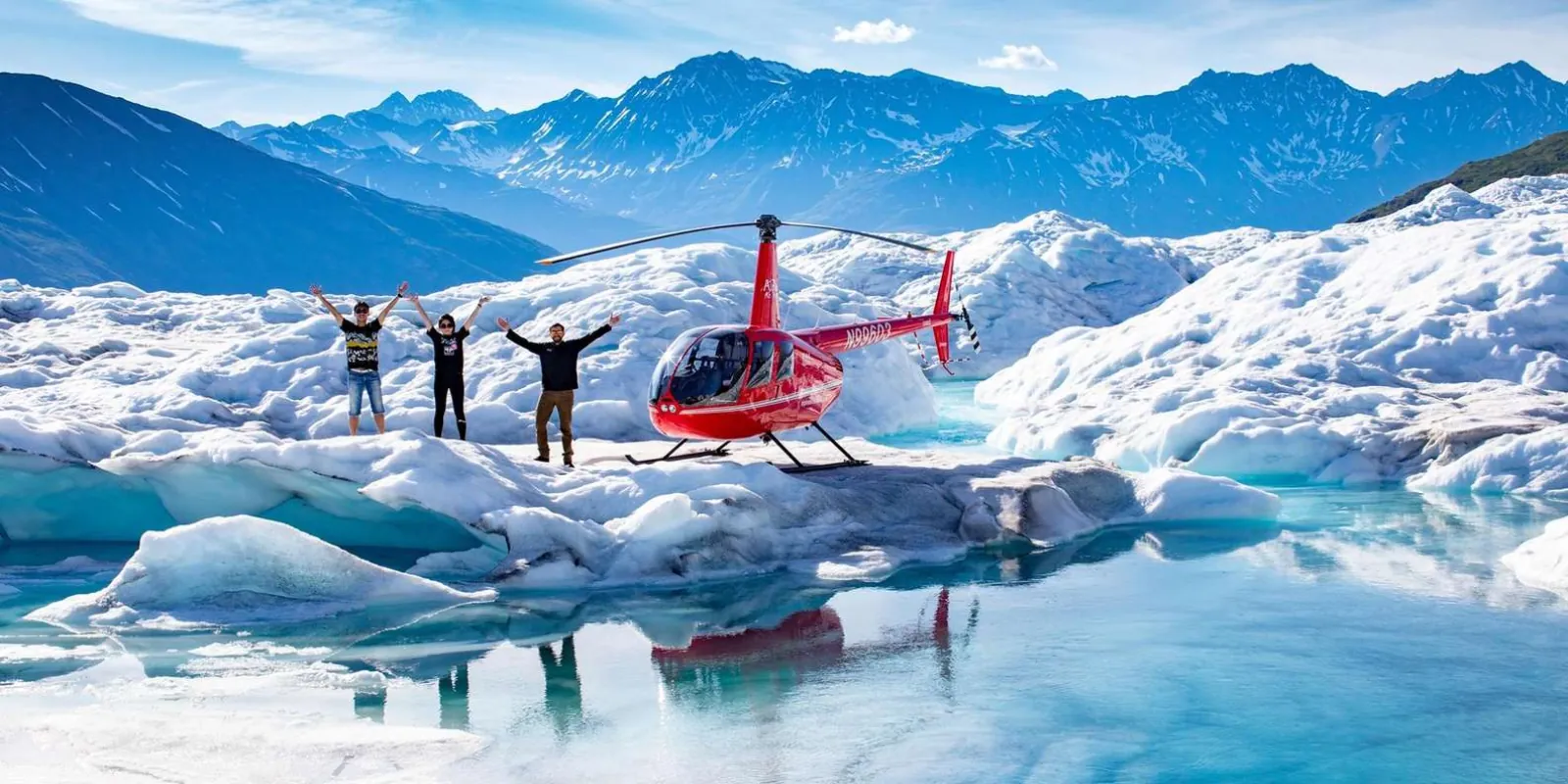 Three guests with arms raised beside a red helicopter parked on the ice, an iceberg-strewn glacier lake in front of them