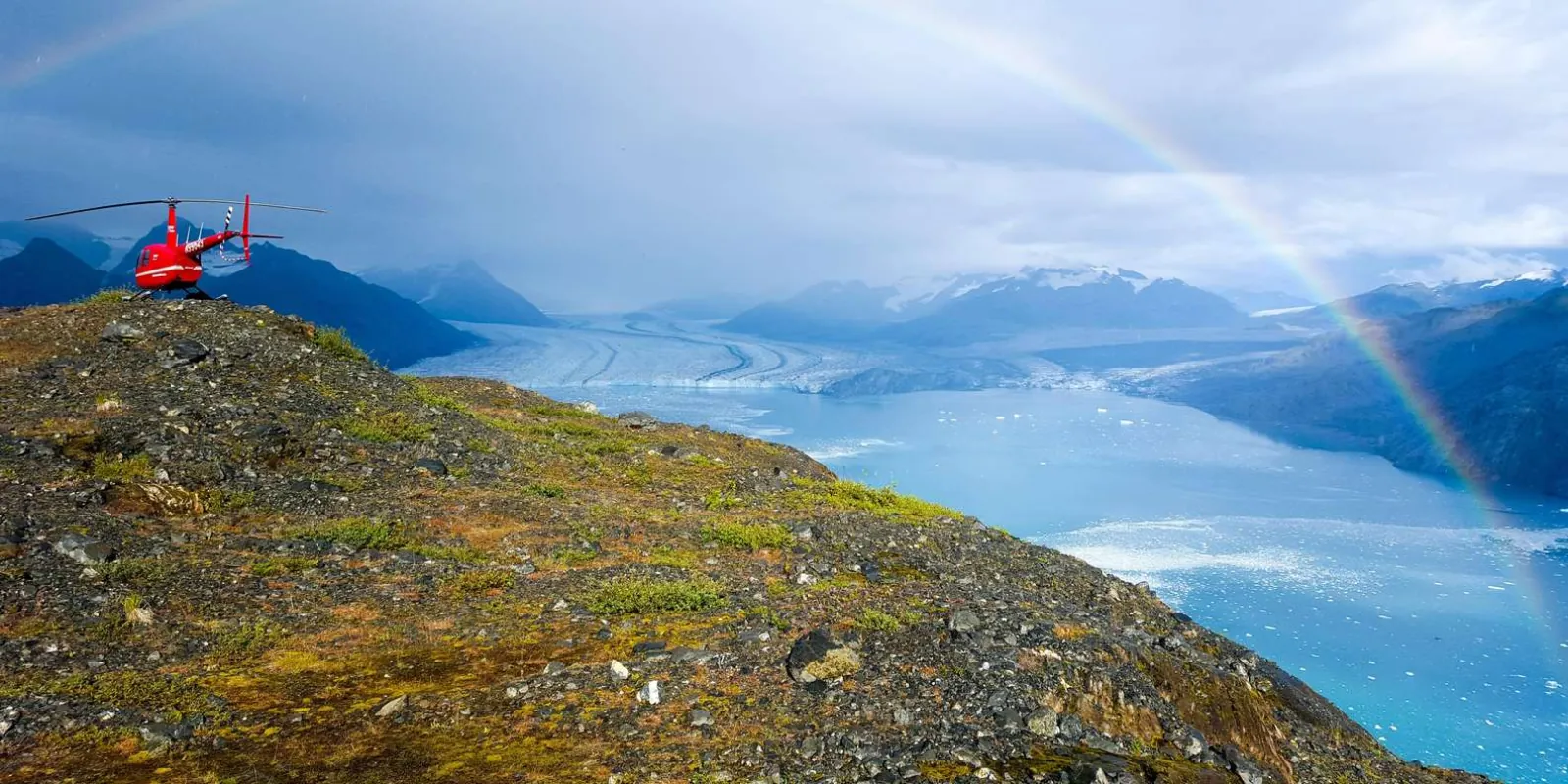 A red helicopter landed on a green alpine ridge above a wide glacier, a rainbow arcing over the valley