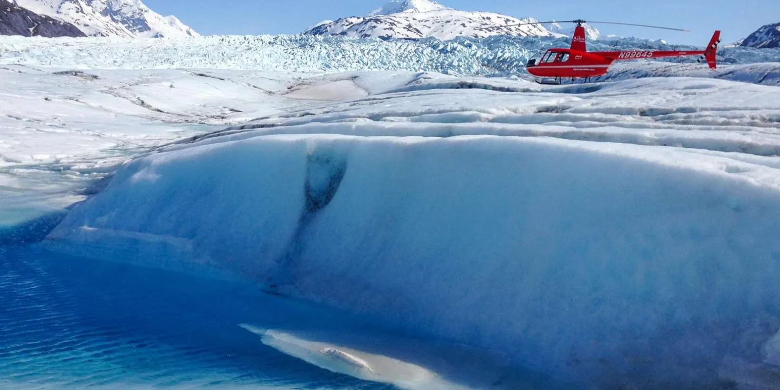 A red helicopter flying low over a smooth blue ice basin at the head of the glacier