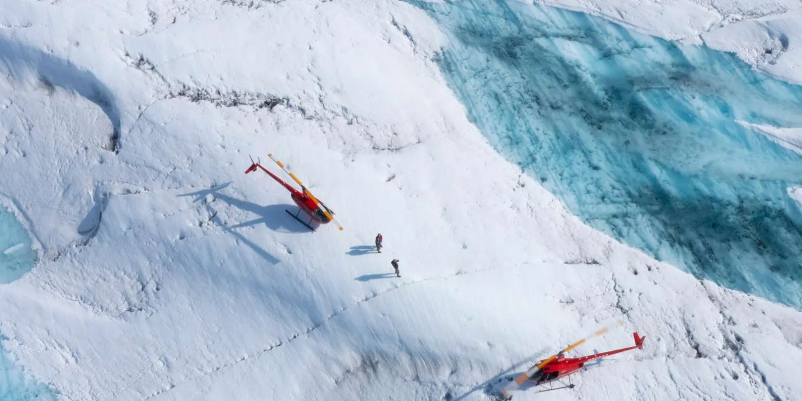 Two red helicopters seen from above on the snow beside a run of deep blue crevasses
