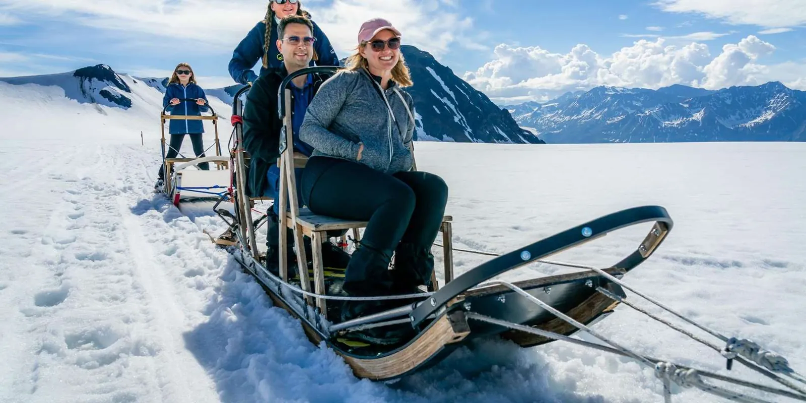 Four guests riding a dog sled across the glacier snowfield with mountains on the horizon