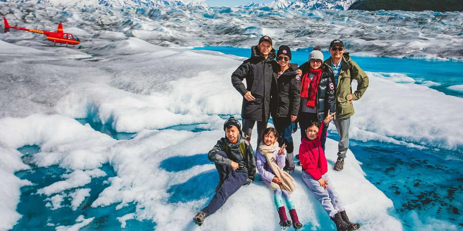 A family of eight posing on the glacier ice beside a blue pool, the red helicopter parked behind them