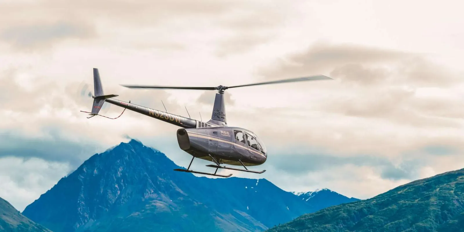 A helicopter in flight above the Chugach Mountains under a soft, high overcast