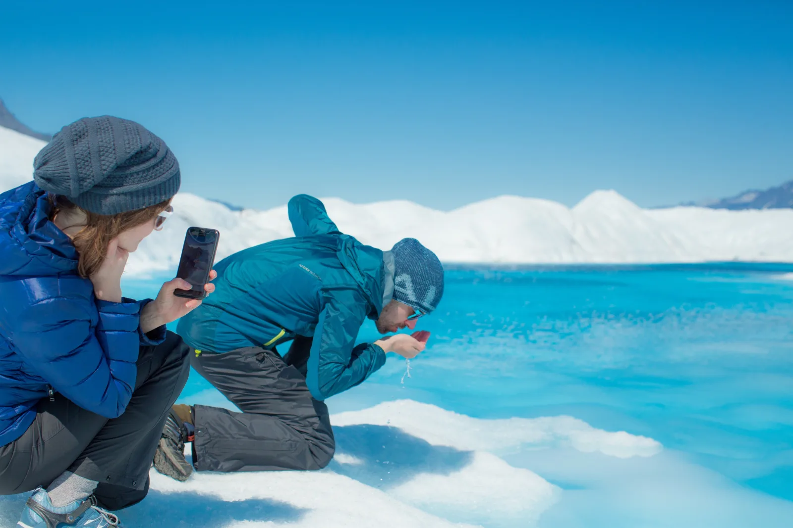 Two guests in blue jackets crouching on the glacier, one drinking meltwater straight from a bright blue pool