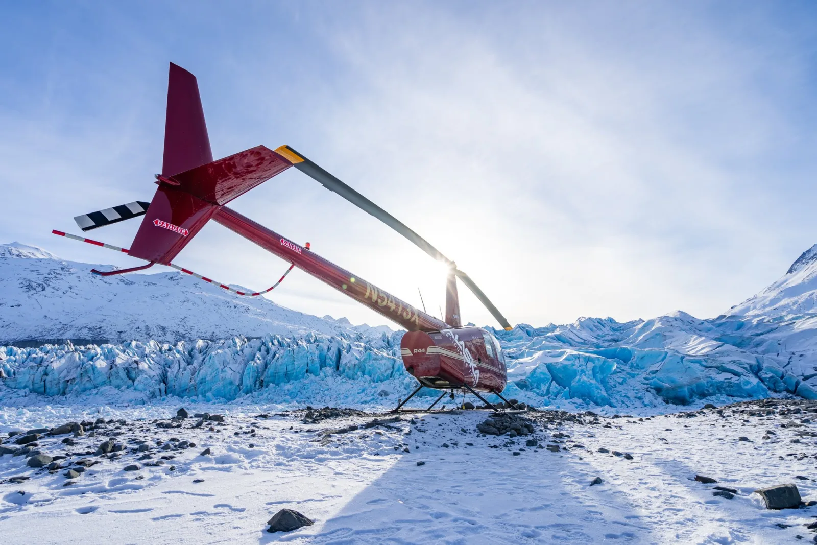 A red helicopter on the glacier with the low Alaskan sun flaring through its rotor blades