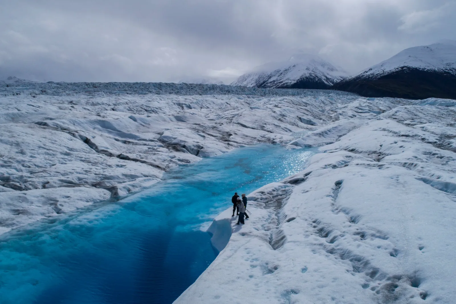 Two tiny figures walking beside a deep blue meltwater stream cut into the white ice of Knik Glacier