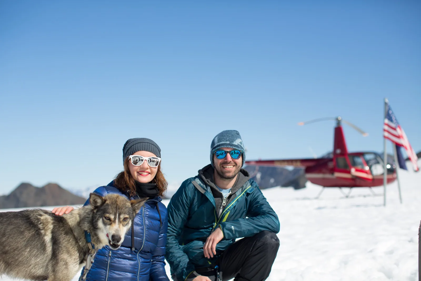 A couple kneeling beside a husky on the snowfield, the red helicopter and an American flag behind them