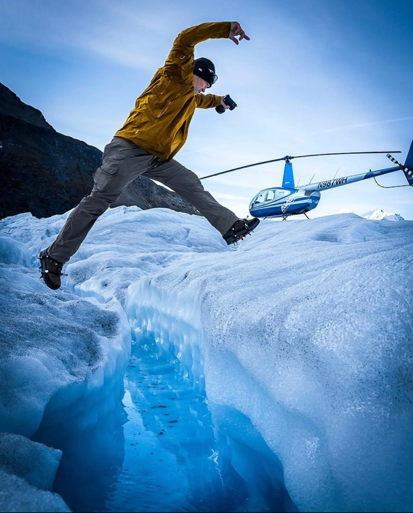 A guest in a yellow jacket leaping across a narrow blue crevasse on the glacier surface