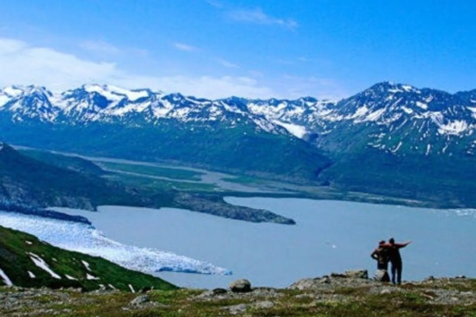 Two hikers on a green slope high above a glacier-fed lake