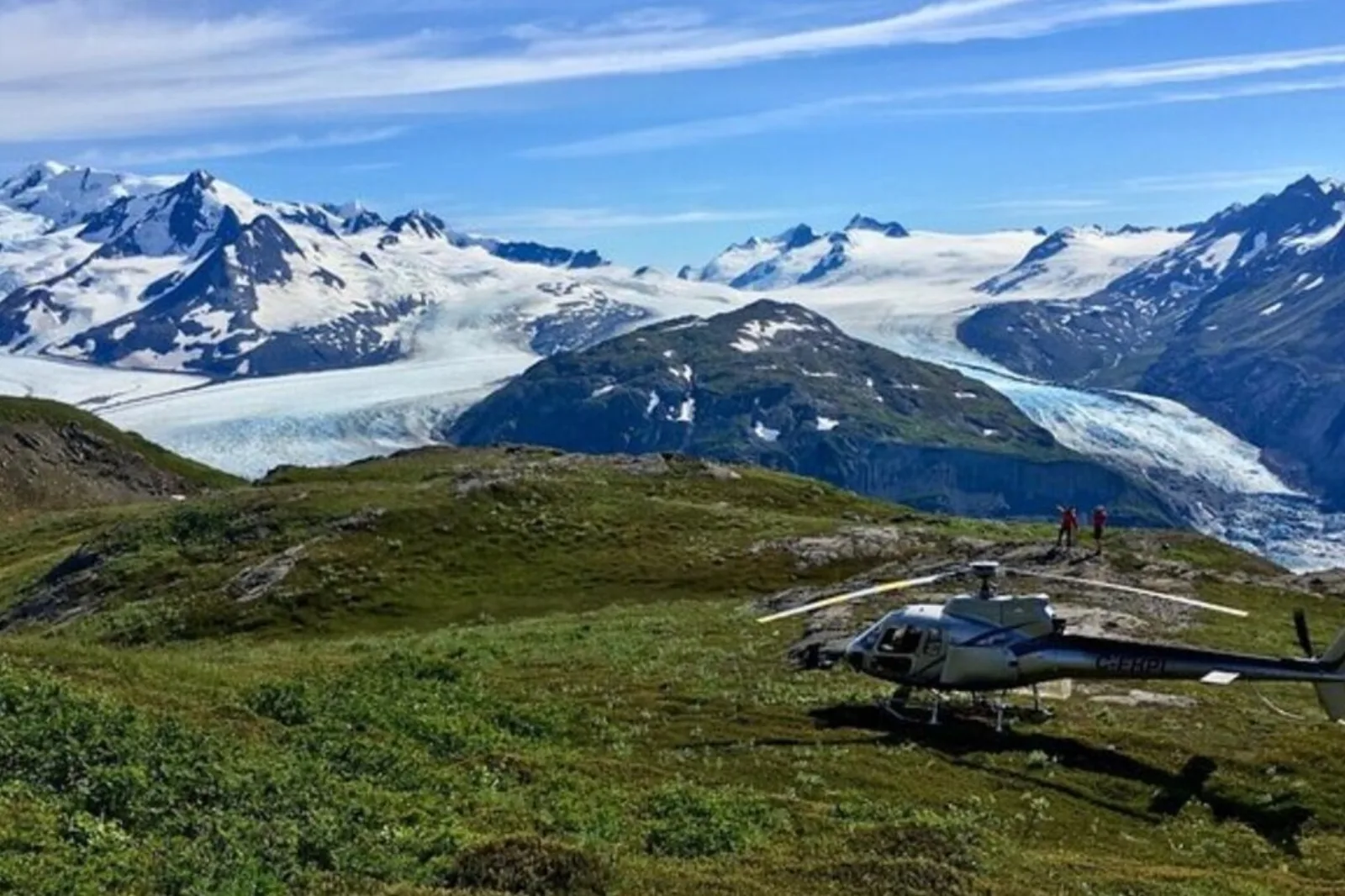 A helicopter parked on a green alpine ridge with the glacier tongue running away into the mountains behind
