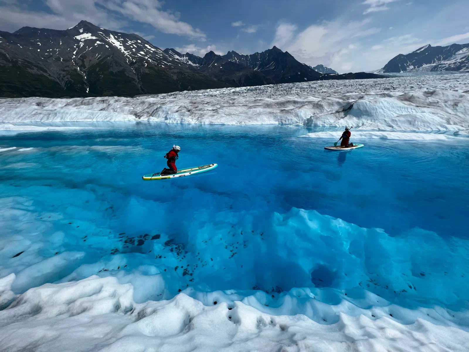 Two paddleboarders crossing a deep blue meltwater lake on the surface of the glacier