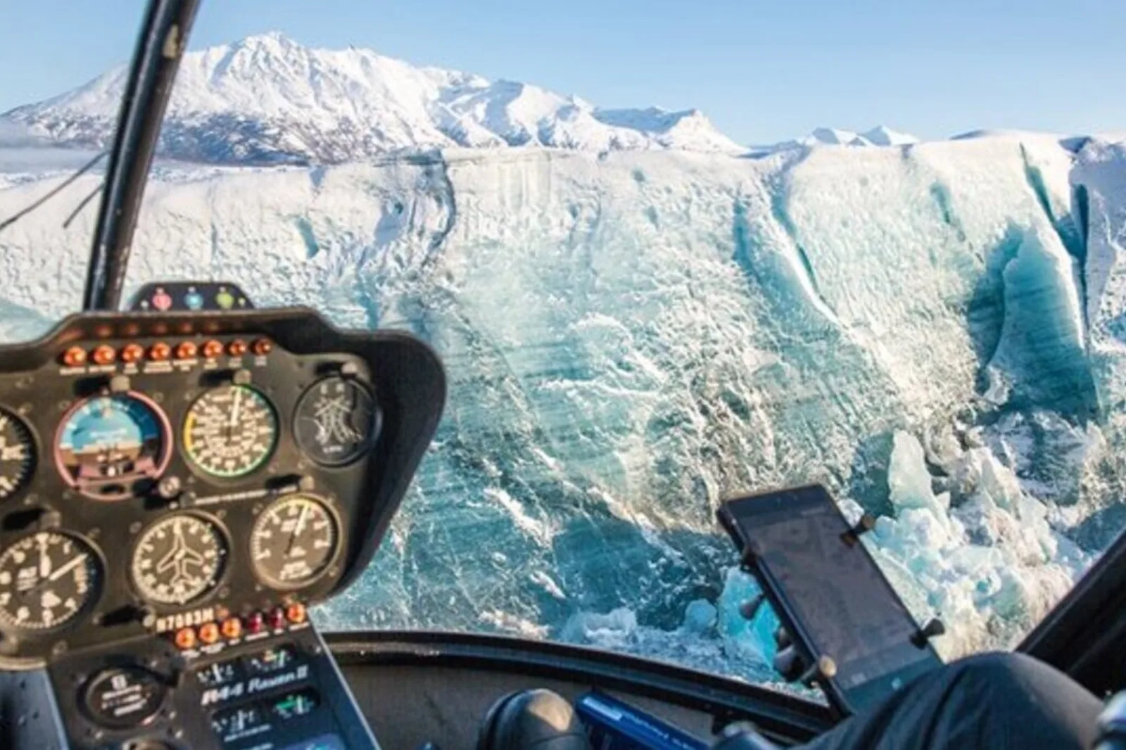 View through the helicopter windscreen towards a towering blue ice wall, the instrument panel lit in the foreground