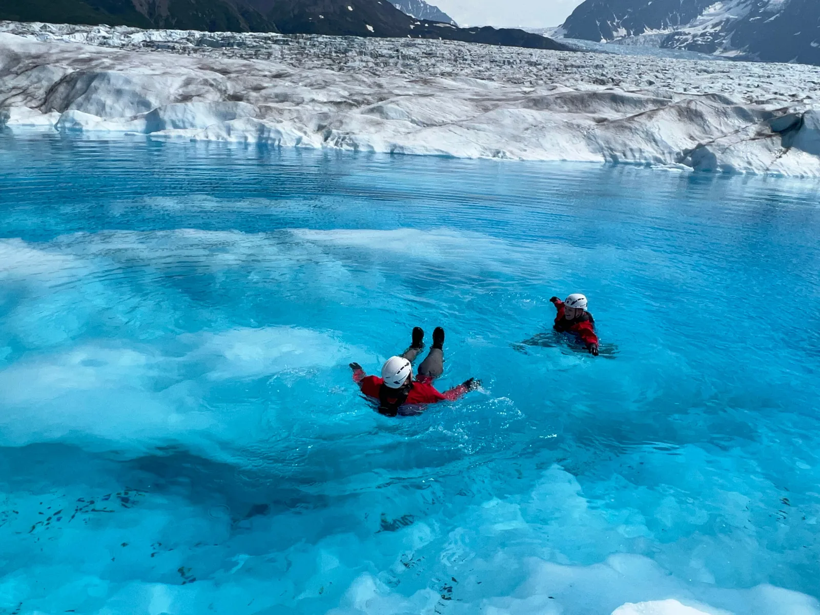 Two guests in drysuits swimming in the bright blue meltwater beneath a glacier ice cliff