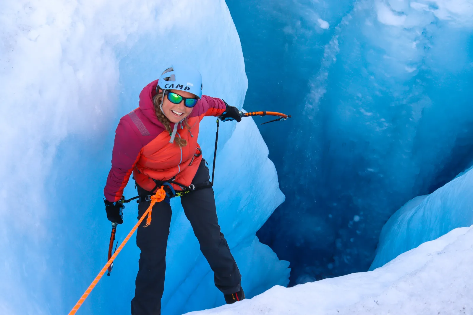A climber in a pink jacket standing at the lip of a blue crevasse with ice tools in both hands