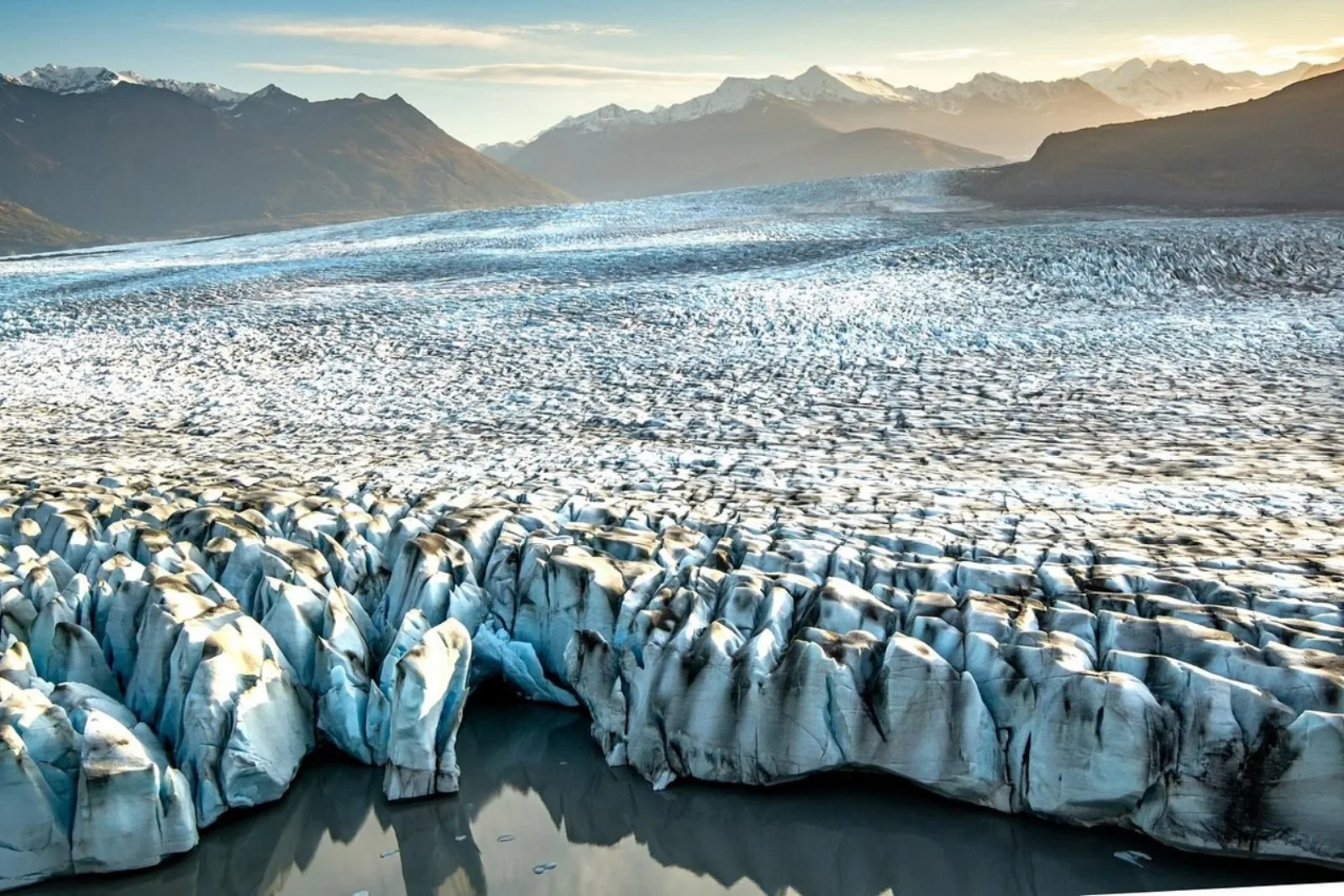 Aerial view of the glacier terminus at low sun, ridges of blue ice meeting the outwash plain